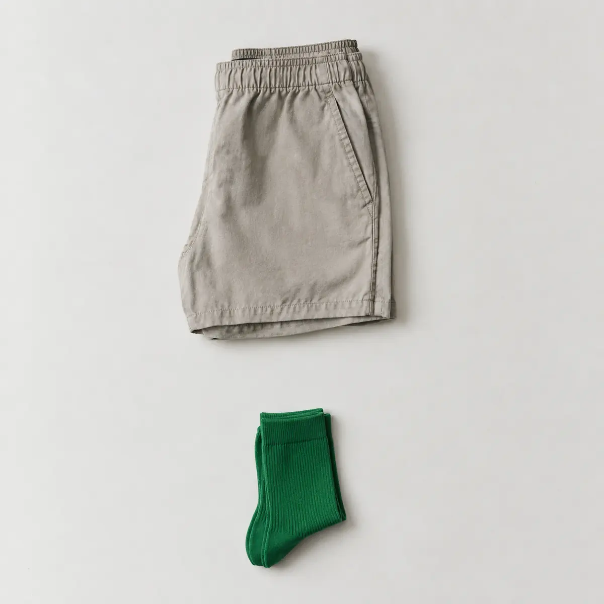 Folded stone coloured cotton shorts on chalk paper with a folded pair of bright green socks placed well below, a wide gap between them.