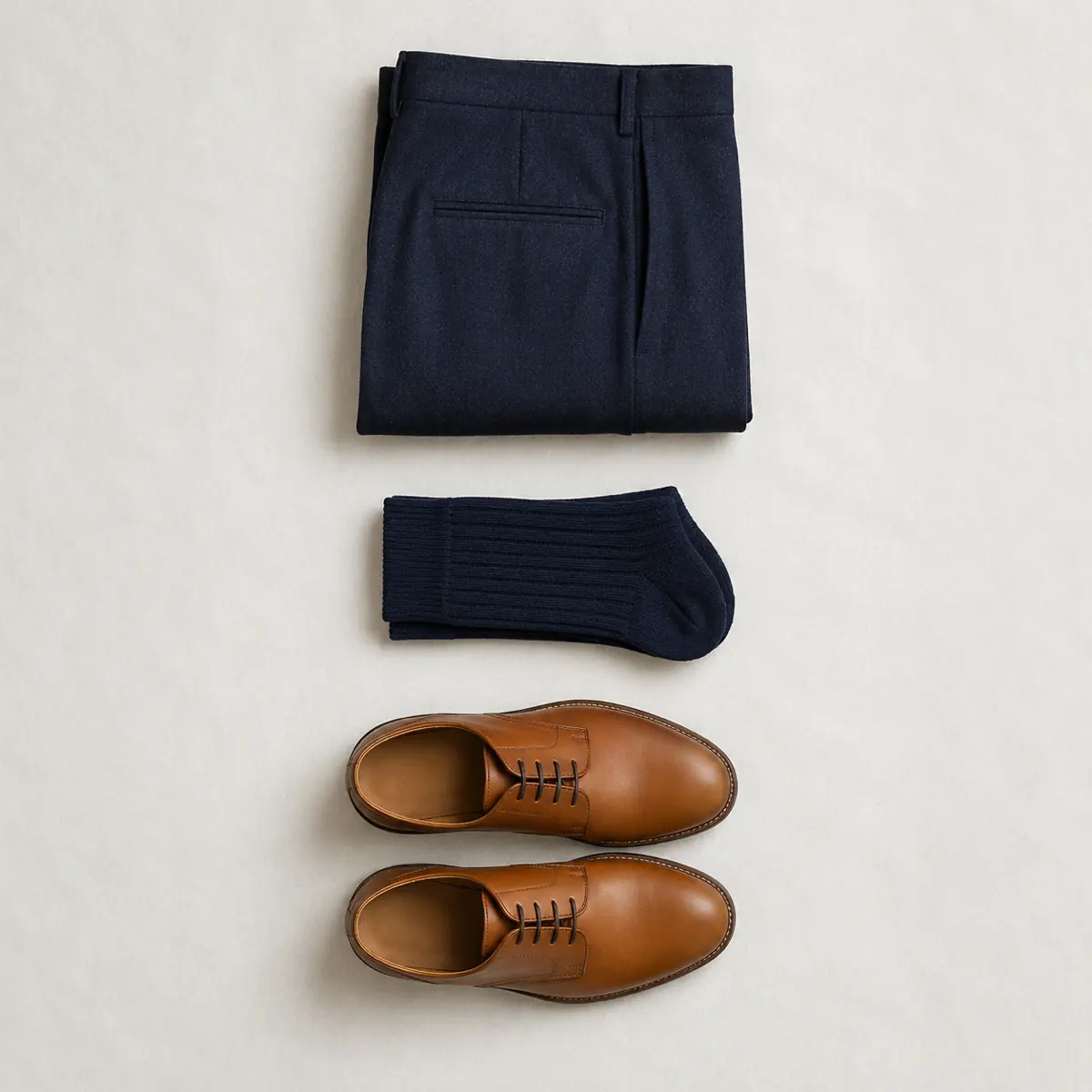 Folded navy wool trousers, folded navy ribbed socks and a pair of tan calf-leather derby shoes laid in one vertical column.