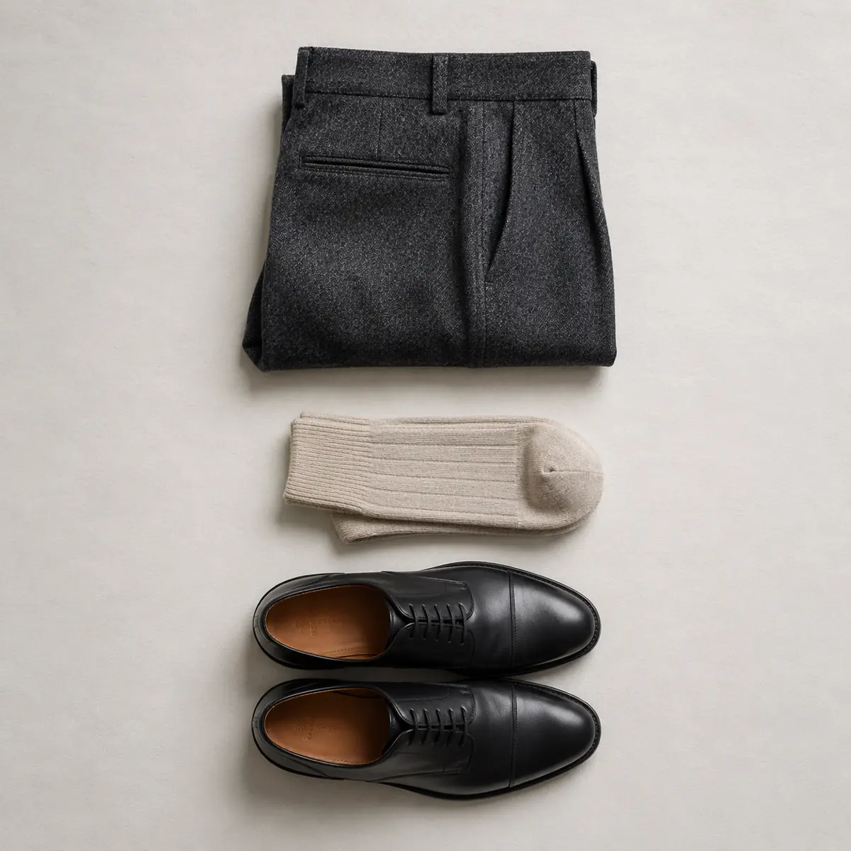 Folded charcoal flannel trousers, folded pale stone cotton socks and a pair of black oxford shoes laid in a vertical column.