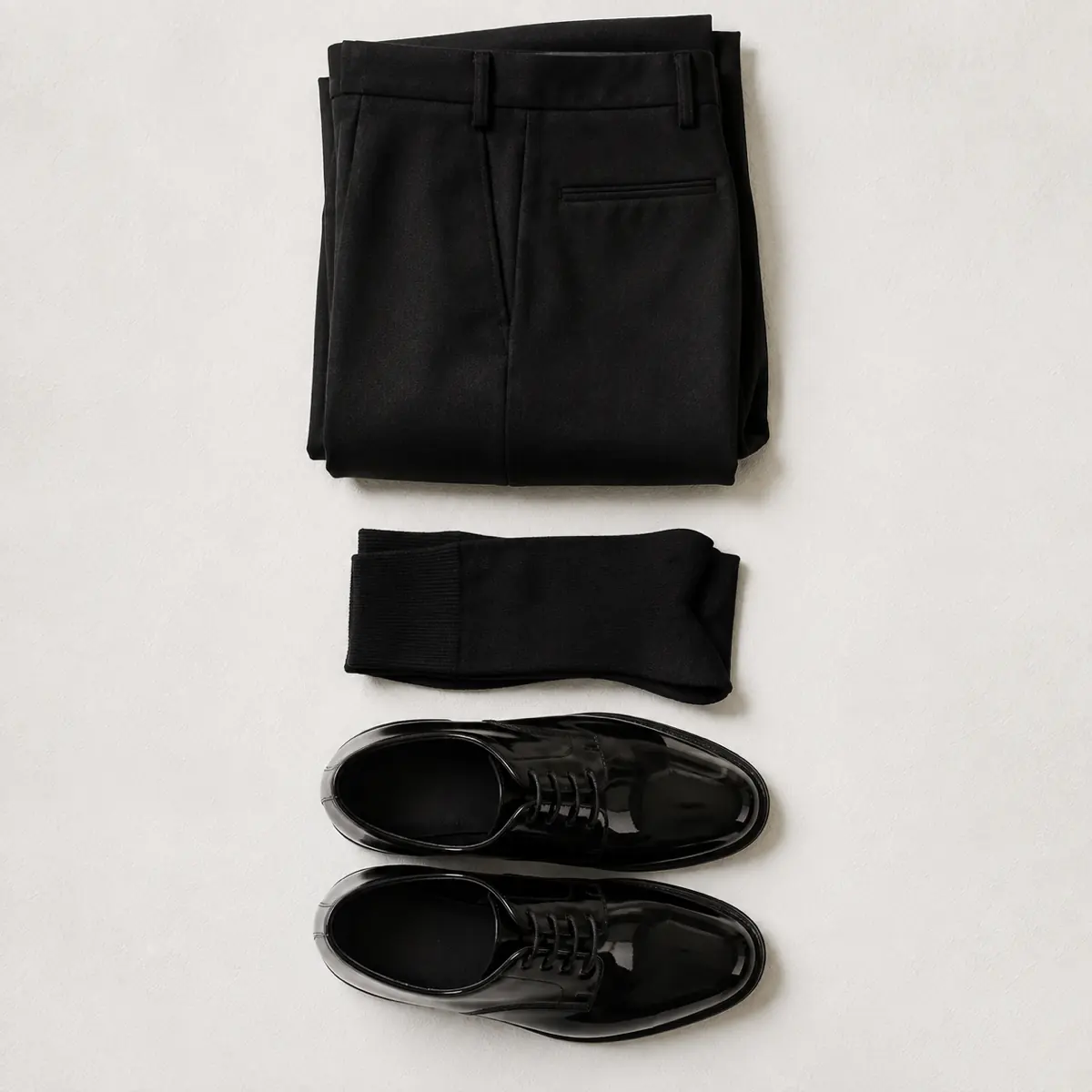 Folded ink black wool trousers, folded ink black fine dress socks and a pair of black patent shoes laid in a vertical column.
