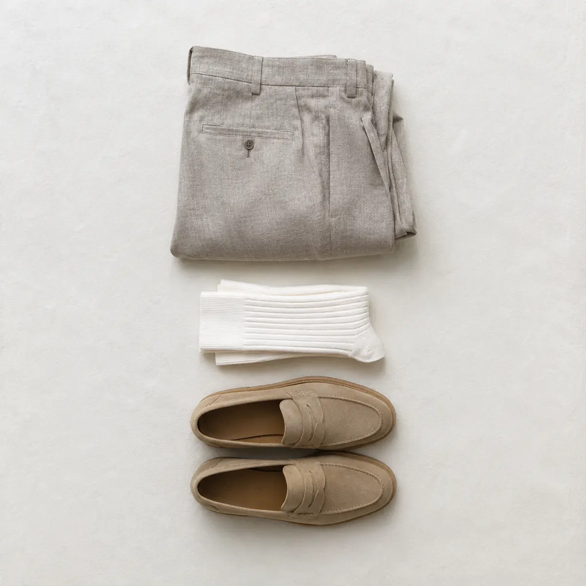 Folded stone linen trousers, folded chalk white ribbed socks and a pair of light tan loafers laid in a vertical column.