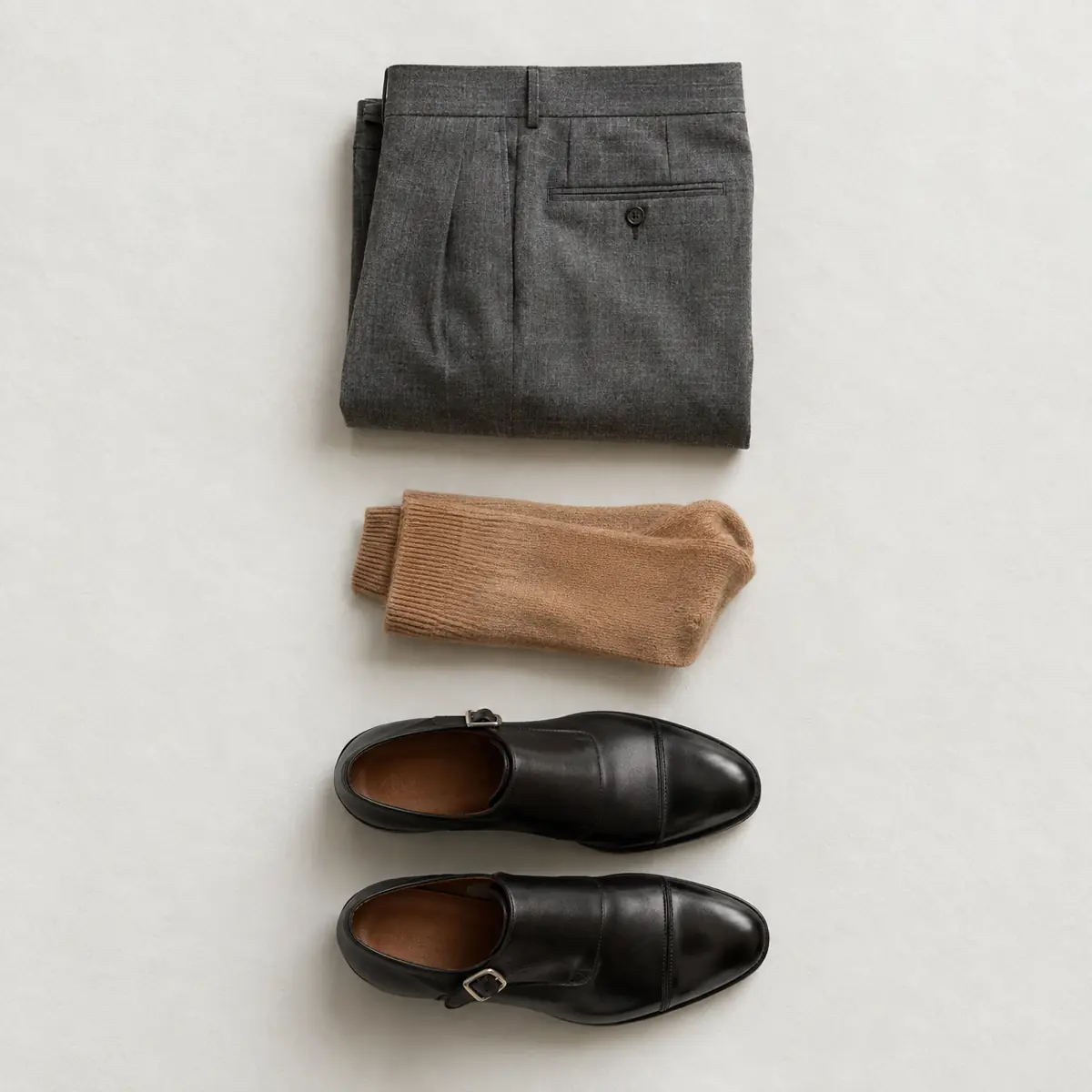 Folded grey worsted trousers, folded camel cashmere socks and a pair of black leather monk shoes in a vertical column.