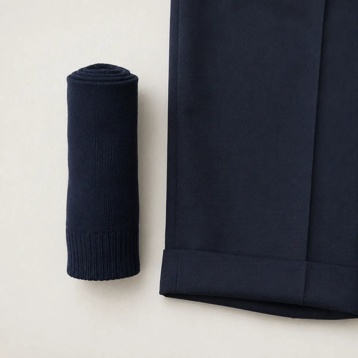 A rolled pair of navy over-the-calf socks standing on end beside a folded navy trouser, seen from directly above on chalk paper.