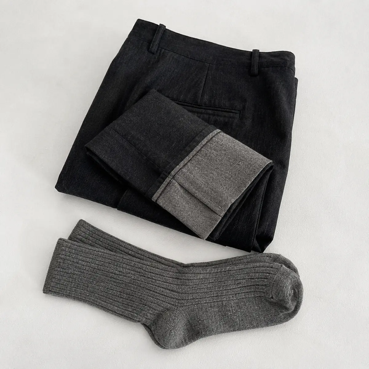 Folded dark charcoal trousers with the hem turned back, laid flat with a pair of grey socks below.
