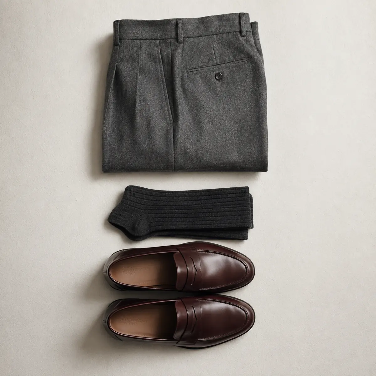Folded mid grey flannel trousers, folded charcoal socks and a pair of oxblood leather loafers in a vertical column.