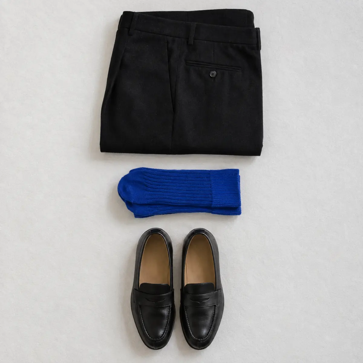 Folded black wool trousers with folded bright cobalt blue socks and a pair of black leather loafers in a vertical column.