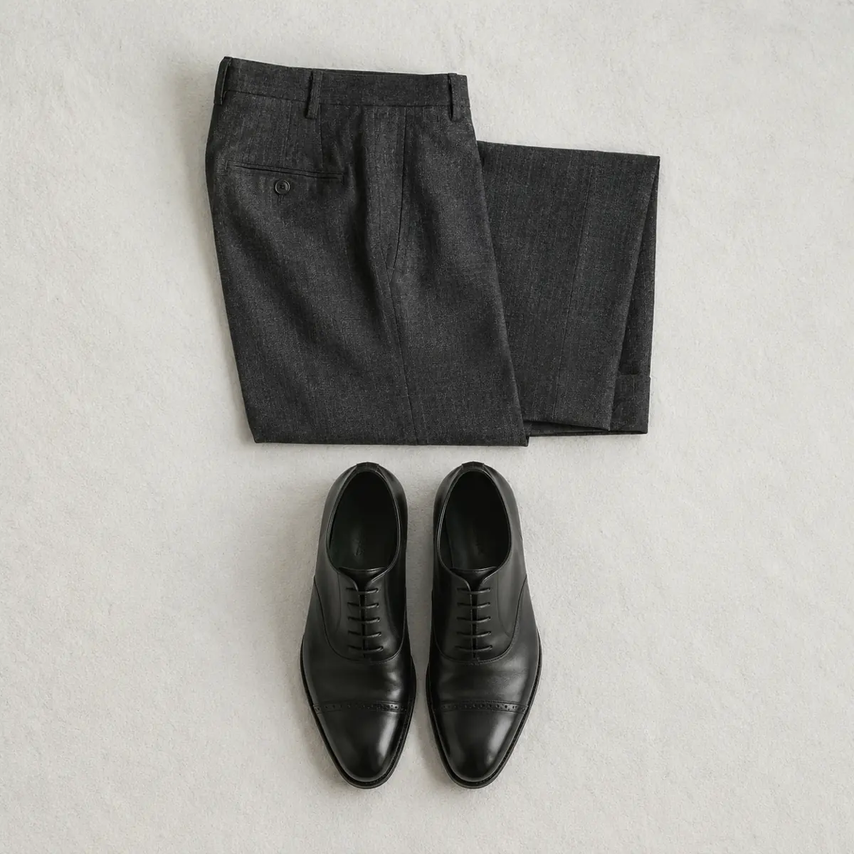 Folded charcoal trousers with the hem laid open flat and a pair of black oxford shoes placed below, with no sock between them.