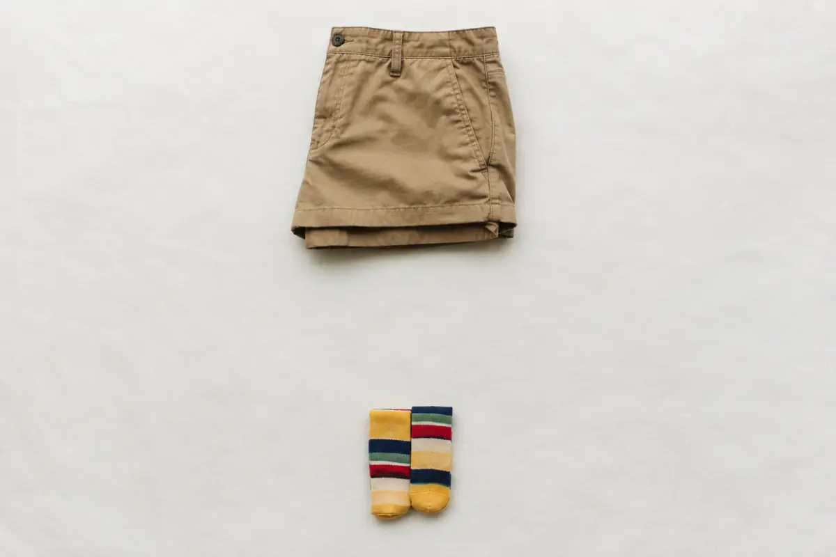 Folded khaki cotton shorts alone on chalk paper with a folded pair of bright striped socks placed far below, a wide gap between them.