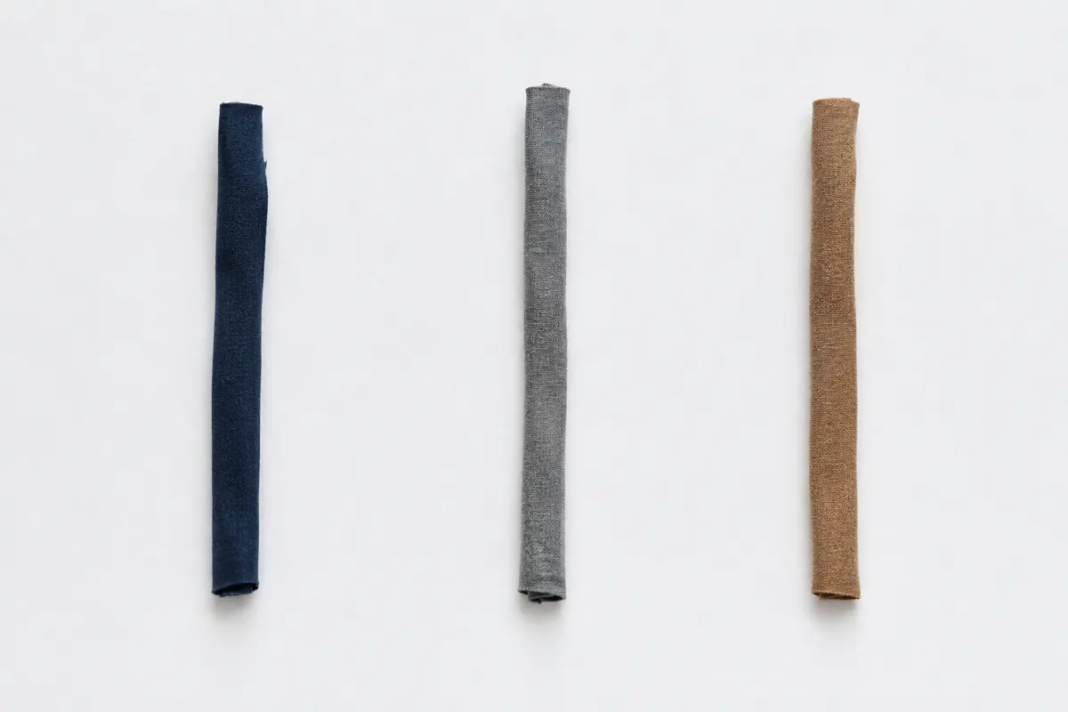 Three narrow folded fabric strips in navy, grey and camel laid far apart as three thin vertical lines on a wide empty sheet.