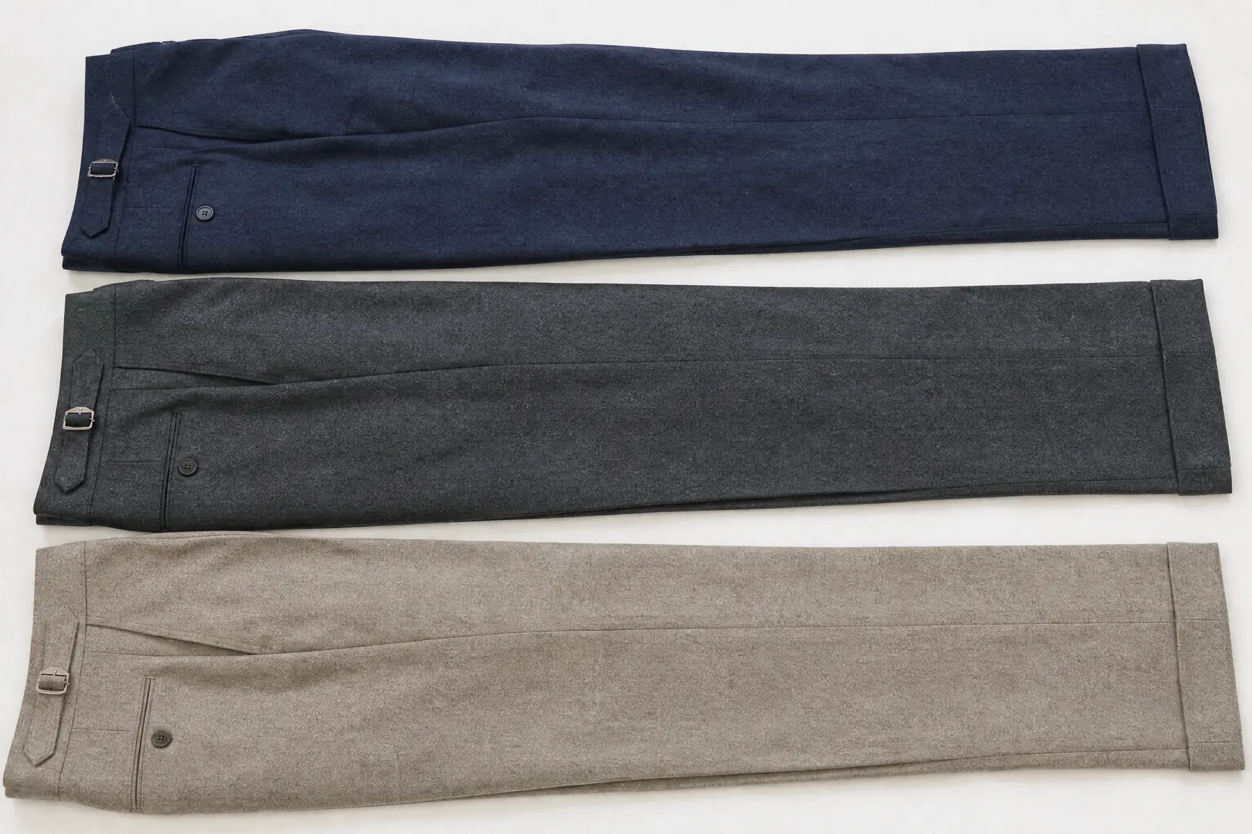 A wide overhead composition of three folded wool trouser lengths in navy, charcoal and stone laid as three long parallel bands.