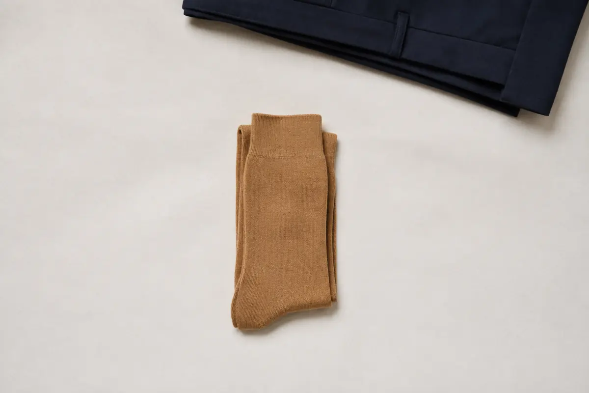 A single folded pair of camel socks alone at the centre of a large sheet of chalk paper, with a folded navy trouser entering from the top edge.
